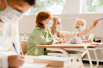 Teacher explaining helping her student pupil schoolgirl with difficult task topic module test wearing protective face masks against Covid19 coronavirus. Pandemic at school concept