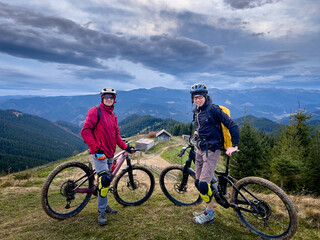 Fototapeta premium Two cyclists men riding electric bikes outdoors. Portrait of male tourists resting on the top of hill, enjoying beautiful mountain landscape in the cloudy evening. Concept of active leisure.