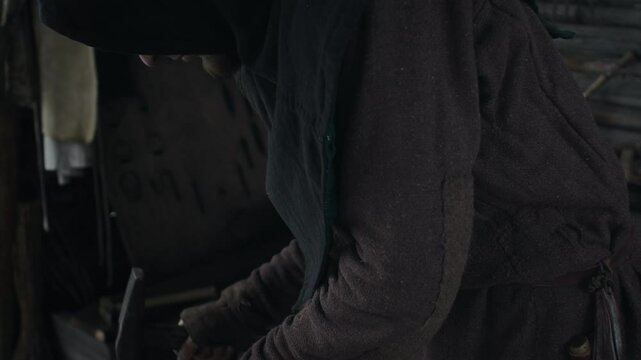 Medium close-up tilt shot of focused young male blacksmith in hooded cloak and peasant coat holding piece of hot iron with tongs and forging it with hammer on anvil, while doing hard labour in smithy