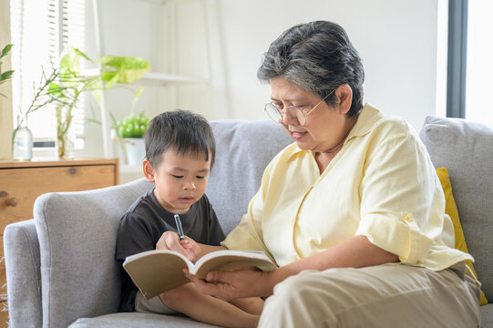 Senior Asian grandmother teaching writing and relaxing with her grandchild at living room happily. Diversity multi generation family activity lifestyle concept. Asian people domestic lifestyle concept