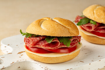 Close up of sandwich or panino with sliced salami, sausage, tomato, cheese, rocket or arugula on a white wooden board.