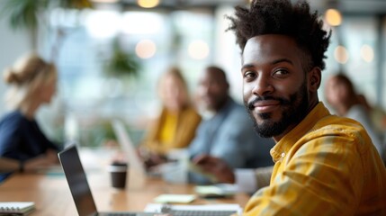 A man in a yellow shirt sits at a table during a meeting with colleagues in a modern office. He looks towards the camera, exuding confidence and approachability.