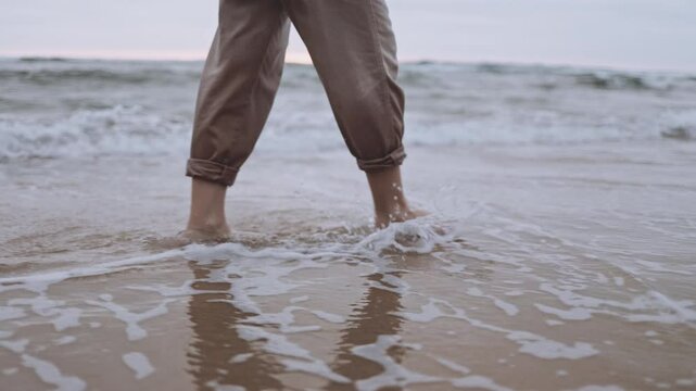 An unrecognizable woman walks alone on tranquil beach at sunset. The calm ocean waves wet the sand and her feet. The cinematic scene captures the melancholy and reflective mood of a summer