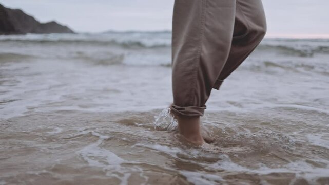 An unrecognizable woman walks alone on tranquil beach at sunset. The calm ocean waves wet the sand and her feet. The cinematic scene captures the melancholy and reflective mood of a summer