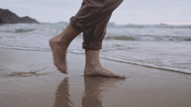 An unrecognizable woman walks alone on tranquil beach at sunset. The calm ocean waves wet the sand and her feet. The cinematic scene captures the melancholy and reflective mood of a summer