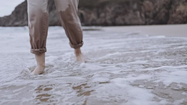 An unrecognizable woman walks alone on tranquil beach at sunset. The calm ocean waves wet the sand and her feet. The cinematic scene captures the melancholy and reflective mood of a summer