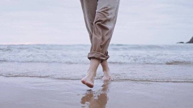 An unrecognizable woman walks alone on tranquil beach at sunset. The calm ocean waves wet the sand and her feet. The cinematic scene captures the melancholy and reflective mood of a summer 
