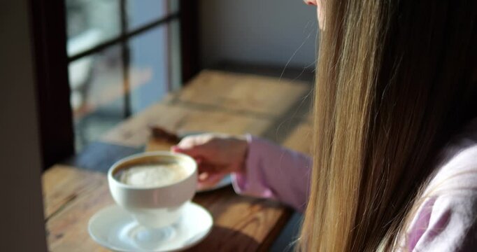 Woman with long loose hair drinks hot coffee sitting near coffee shop window at sunshine closeup. Female refreshes with decaf beverage at wooden table