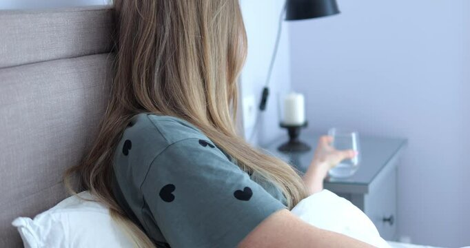 Tired woman with long hair drinks water from glass resting in bedroom slow motion. Sick lady refreshes with clear beverage following doctor recommendation in bed