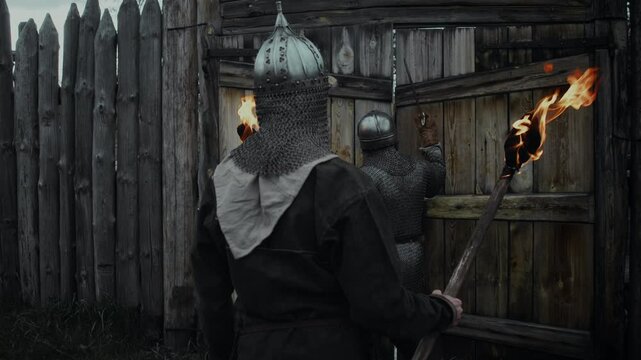 Medium rear follow shot of two medieval warriors in chainmail, helmets, carrying burning torches, approaching fortress while returning from patrol, banging on gate, then greeting guard and walking in