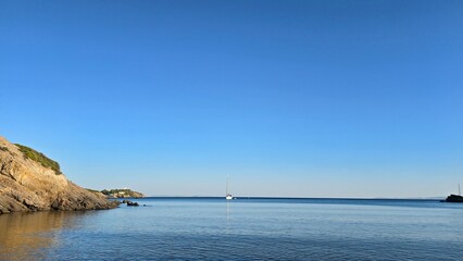 Sea view from Bilali Beach on Oinousses Island, Greece. The scene features lush forest on the sides, a vibrant blue sea, a boat in the water, and a glimpse of Pateroniso in the distance