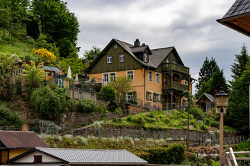 A house with a green roof sits on a hillside. The house is surrounded by a lush green garden and has a large patio area. The sky is cloudy, giving the scene a somewhat moody atmosphere