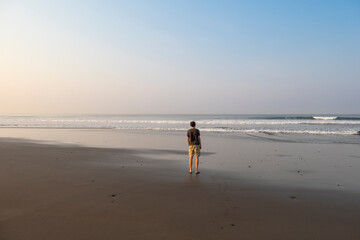 Young fit man in shorts and t-shirt standing on the beach looking at crushing waves at early morning sunrise hour. La Libertad, El Salvador.