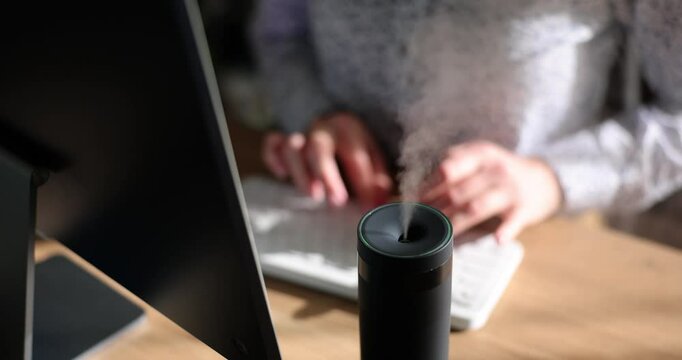 Woman types on keyboard of computer at wooden table while diffuser spreading fragrance. Freelancer works at home enjoying pleasant aroma slow motion