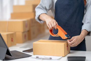 Person sealing a cardboard box with tape in a fulfillment center, surrounded by packages, laptop, and clipboard.