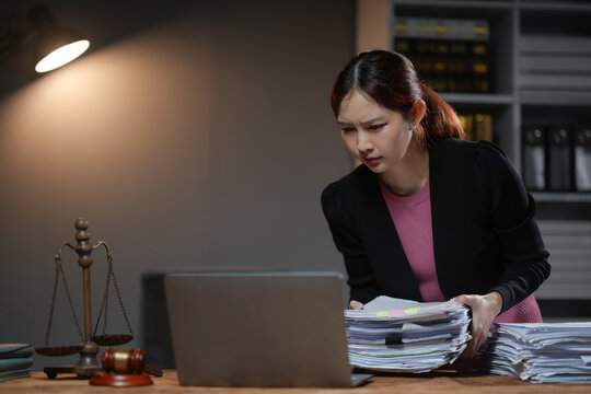 Businesswoman working on a laptop in an office environment, focused on paperwork and legal documents under a desk lamp light.