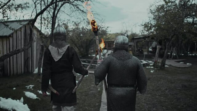 Medium rear follow shot of two knights in chainmail armour and helmets walking through garrison yard, carrying burning torches, past wooden huts, in spring evening, snow lying on ground