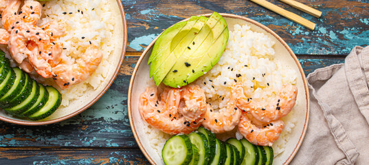 Two white ceramic bowls with rice, shrimps, avocado, vegetables and sesame seeds and chopsticks on colourful rustic wooden background top view. Healthy asian style poke bowl.