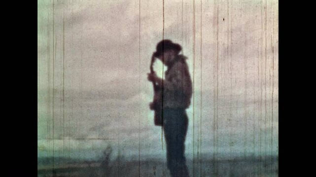 Man playing guitar, singing on cloudy sky background in wild field nature. Male cowboy in hat enjoy playing musical instrument in summer outdoors. Vintage color film. Travel archive. Retro 1960s