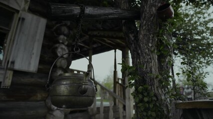 Close-up shot of copper teapot hanging on chain off tree branch, in front of wooden peasant hut, in medieval village or historical educational park on cloudy day