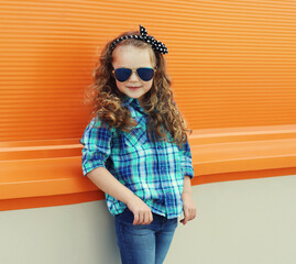 Happy kid girl with curly hair, stylish little child posing in the city on orange wall