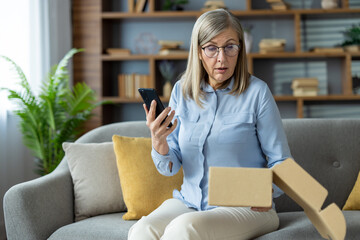 Senior woman experiencing surprise and confusion while unboxing unexpected package and using smartphone, sitting on sofa in living room. Green plant and bookshelf in background.