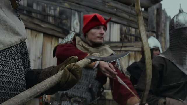 Medium shot of young Caucasian male archer in red uniform and armoured breastplate drawing bow and aiming arrow at advancing enemy, while defending wooden fortress with companions