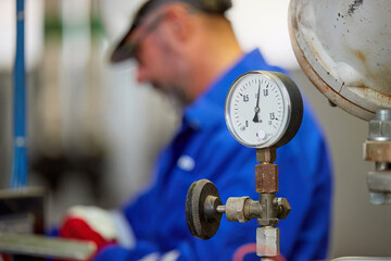 Close-up of pressure gauge in a modern boiler room with an engineer working in the background, wearing a blue uniform and safety gear. Industrial setting with focus on equipment and maintenance.