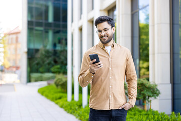 Hispanic man in casual attire standing outside modern office building using smartphone. Daylight, urban setting, technology and business concepts.