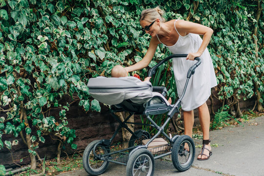 Woman in sundress talking calmly to infant while pushing stroller along sidewalk on green street