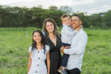 latin family posing happily in a green field, all four are smiling.
