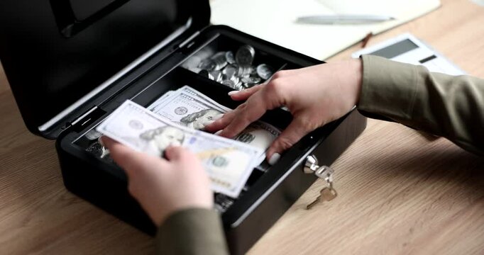 Woman takes dollar bills out of cash register of accountant with safety lock and key. Black box with banknotes and coins on wooden table slow motion
