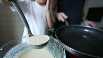 Close-up of child pouring pancake batter into pan, kitchen activity, child focused on task, parent assisting, white t-shirt, morning light, home environment, making breakfast together