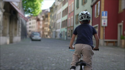 Obraz premium Child riding bicycle with helmet on cobblestone street in charming old town with colorful buildings, promoting outdoor activity and exploration in historic urban environment