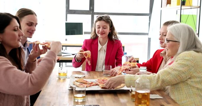Delighted colleagues take slices of delicious pizza sitting at big wooden table in premise. Happy man and women enjoy lunch at work break in office slow motion