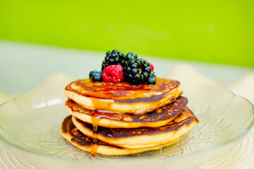 Imagen horizontal de un plato de hot cakes o panqueques con frutos rojos y miel en una mesa con fondo verde