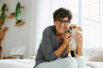Smiling mature woman with Chihuahua dog looking at camera at home