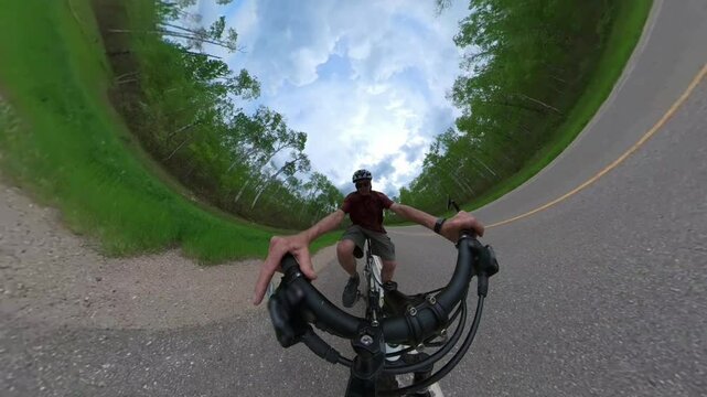 Extreme wide-angle view from a 360 degree camera looking at a male cycler pedalling along the side of a paved road that runs through a green forest.  
