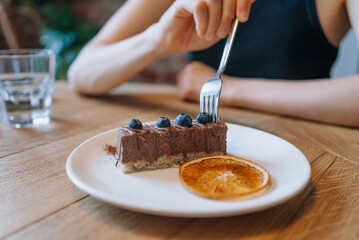 Woman hand holding fork picking up slice of chocolate cake topped with blueberries and garnished with dried orange slice, with a glass of water in the background. 