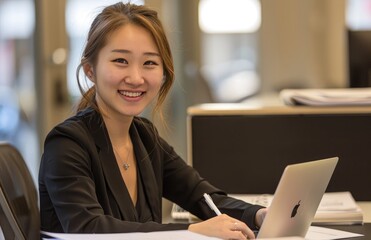 A photo of an Asian woman in professional attire, smiling and working on her laptop at the office desk with documents beside it. The background is blurred to focus attention on the female worker.