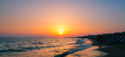 Sunset on the Mediterranean Sea. Natural seascape with calm waves and bright low sun. Side view of the water and beach. The concept of an evening walk. Coastline. Holidays in Italy. Nature wallpaper