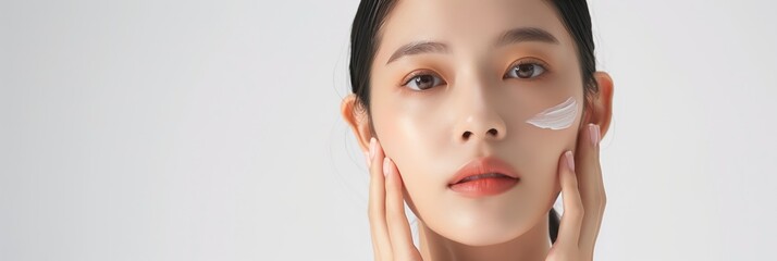 Korean model woman applies cream on face in beauty studio. Close-up shot of face shows gentle eyes, slightly open mouth. Wears neutral gray shirt and white makeup, pink lip color.