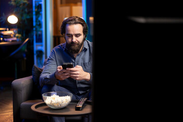 Man enjoying bowl of popcorn while watching cable programs on TV and texting friends on phone. Spectator on couch in living room eats snacks and talks with mates on smartphone