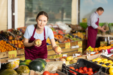 Polite friendly saleswoman standing at counters in greengrocery store, holding ripe red apples...