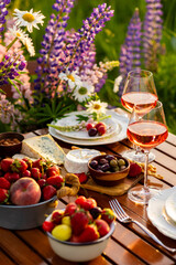 Romantic table decor for a loving couple on the blooming meadow with purple lupines. Two glasses of wine, flowers in a vase, silverware, fruits, wooden furniture and picnic basket. Sunset, golden hour