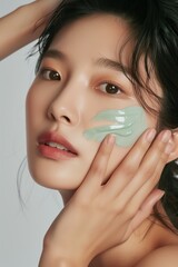 Korean model woman applies green facial mask on face. Close-up view of beauty studio promo with white background. Model gently holds small amount of cream in hand. Focus on details of face, hands.