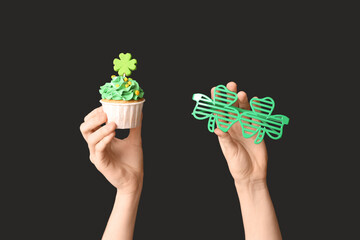 Female hands with tasty cupcakes and plastic eyeglasses for St. Patrick's Day on black background