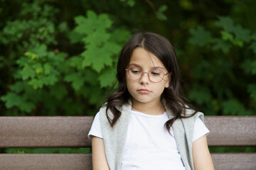 A little sad schoolgirl girl with glasses sits on a bench. Difficult childhood concept