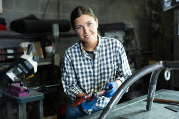 Confident young female worker in casual checkered shirt and gloves, standing in metal workshop with...