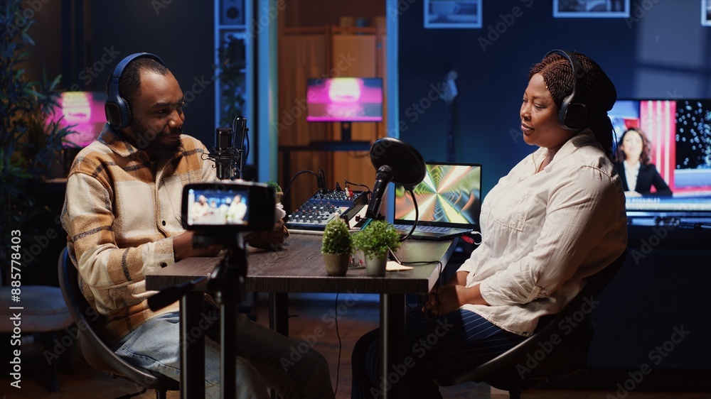 Poster radio show presenter using professional camera to record live conversation with man in personal stud